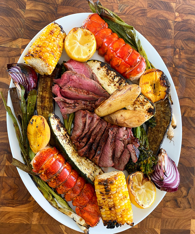 A grilled surf-and-turf platter by Chef Olintho: lobster tails, sliced steak, charred corn, and grilled lemons
