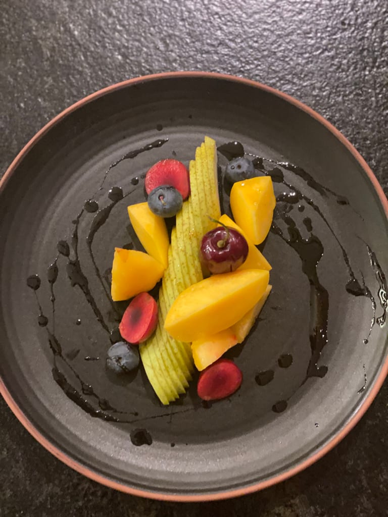 A plated fruit course by Chef Olintho: sliced mango, fanned pear, berries, and a cherry over a dark drizzle on a dark ceramic plate